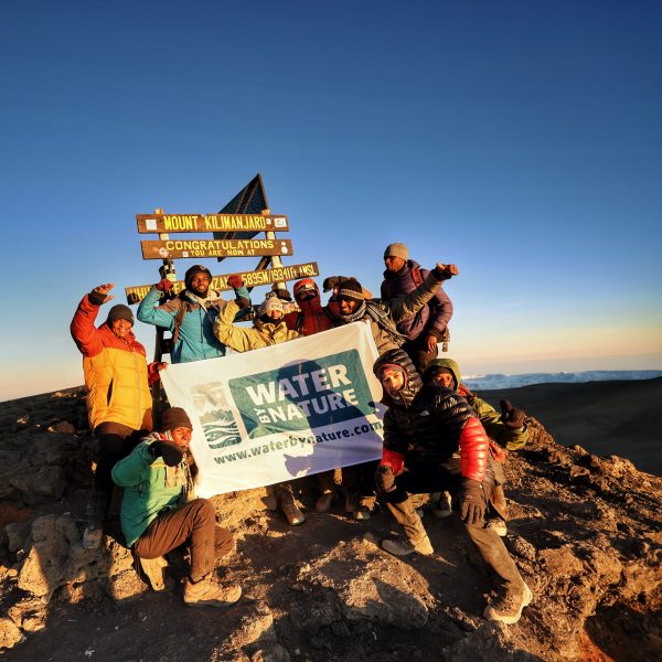 A group of porters and guests standing at Uhuru peak at the top of Kilimanjaro holding a water by nature flag