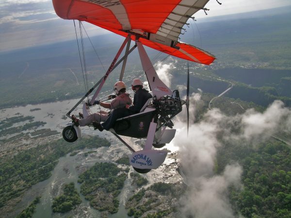 A microlight with orange wing flies high above the batoka gorge and Victoria Falls, with water spray from the falls forming a cloud