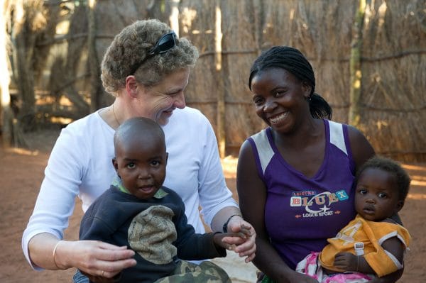 Two women - one black and one white - holding infants on their laps in an African village setting