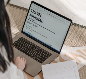 Girl looking at computer screen that says travel journal on it with a paper diary alongside